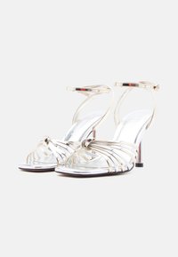 Ζευγάρι μεταλλικών χρυσών σανδαλιών με λουράκια στον αστράγαλο και ανοιχτά δάχτυλα, με ψηλό τακούνι, σε λευκό φόντο.