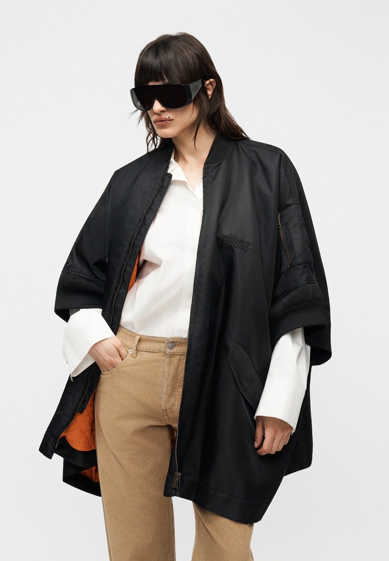 Femme portant une cape noire surdimensionnée à fermeture éclair sur une chemise blanche et un pantalon beige, avec de grandes lunettes de soleil noires, sur un fond uni.