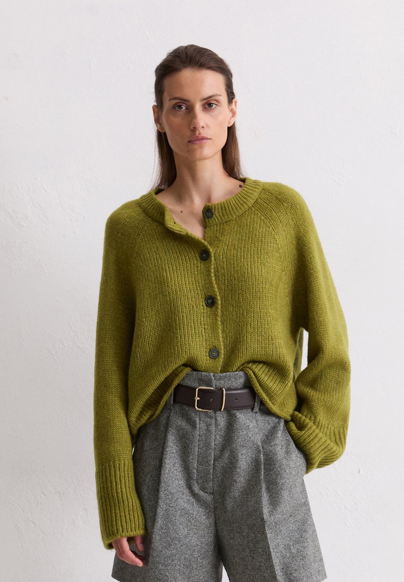 Cardigan en tricot vert avec un col rond et cinq boutons, associé à un short plissé gris et une ceinture marron foncé. Tissu texturé.