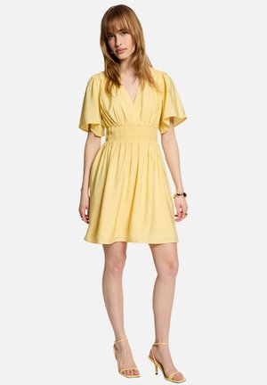 Kvinde iført en gul knælængde kjole med pufærmer, V-udskæring, smock-talje, gule hæle med stropper og gyldne armbånd på sit højre håndled.