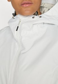 Persoon in een wit hoodie-jack, tot aan de hals dichtgeritst, met verstelbare trekkoorden zichtbaar rond het capuchongedeelte.