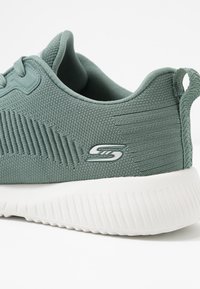 Närbild av en teal-färgad stickad sportskor med en vit tjock struktur i sulan och Skechers-logotyp på sidan.