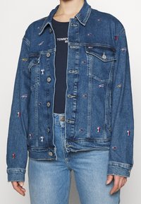 Donkerblauwe spijkerjas met meerkleurige geborduurde logo's en details, voorzien van een knoopsluiting en borstzak. Klassiek kraagontwerp.