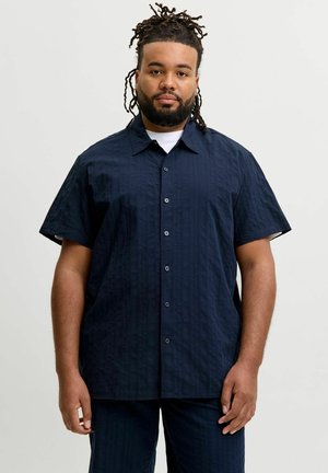 Homme avec des dreadlocks et une barbe portant une chemise bleu foncé à manches courtes boutonnée et un pantalon assorti, debout devant un fond clair uni.