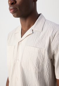 Camisa de manga corta de color beige con textura, dos bolsillos en el pecho y un cuello con muesca, que presenta un sutil patrón de costura y cierre de botones.