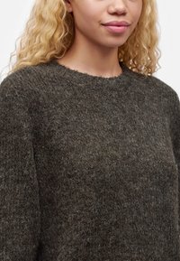 Gros plan sur une personne aux cheveux blonds bouclés portant un pull en maille duveteuse gris foncé avec un col rond.
