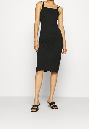 Kobieta ubrana w dopasowaną czarną sukienkę midi z cienkimi podwójnymi ramiączkami oraz czarne plecione sandały na obcasie na jednolitym tle.