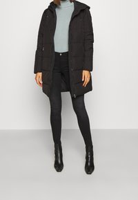 Abrigo de plumas negro con capucha, que presenta un diseño acolchado y cremallera frontal. Lleva puesto un cuello de cisne de rayas en azul claro y jeans negros.