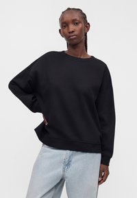 Jeune femme aux cheveux tressés portant un sweat-shirt noir ample et un jean bleu clair sur un fond clair uni.