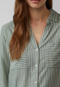 Chemise à rayures vertes à boutons avec col, comportant des rayures verticales blanches et deux poches poitrine. Tissu doux avec une texture lisse.