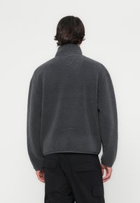 Šedý fleecový pulovr s vysokým límcem, volným střihem, texturovaným materiálem a dlouhými rukávy. Zadní pohled ukazuje jednoduchý design bez ozdob.
