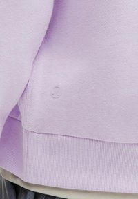 Sweatshirt lilas clair en tissu doux, avec un ourlet et des poignets côtelés, portant un petit logo brodé près du bas.