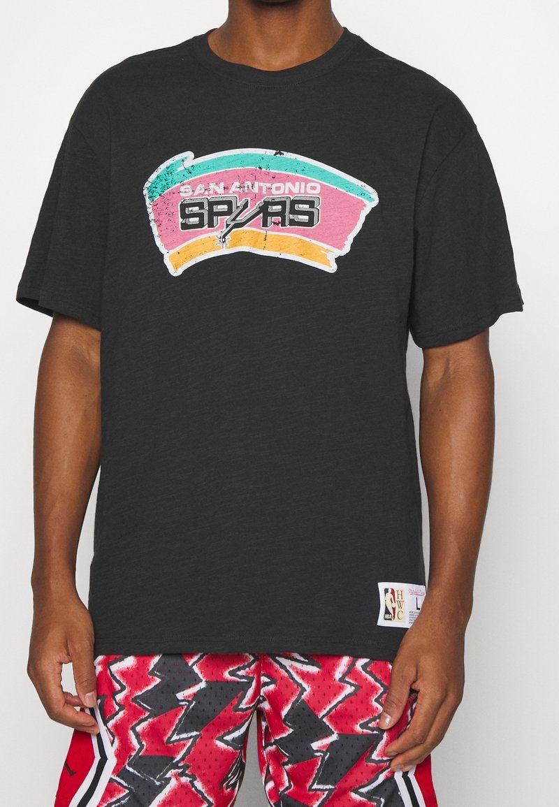 Persona che indossa una maglietta nera dei San Antonio Spurs con logo retrò e pantaloni corti da basket a fantasia rossa su uno sfondo neutro.