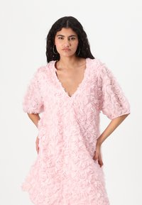 Roze jurk bedekt met gestructureerde, gelaagde rozenvormige applicaties, met een V-hals en korte mouwen, wat een vloeiende silhouteet creëert.