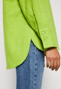Limegrünes, strukturiertes Wollhemd mit abgerundetem Saum, ausgestattet mit knöpfbaren Manschetten und einem lässigen Schnitt, kombiniert mit blauen Jeans.