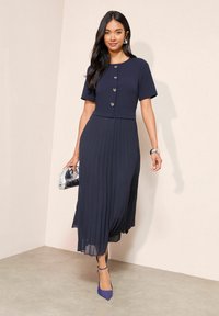 Tengerészkék pliszírozott ruha rövid ujjakkal, szív alakú gombokkal és testhez simuló felsőrésszel. Ezüst kézitáska és lila cipő tartozik hozzá.