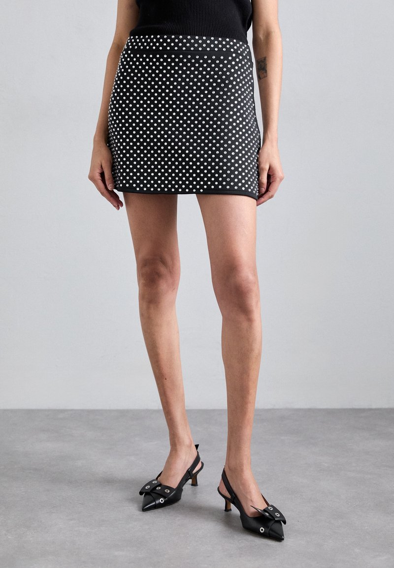 Svart mini-kjole med hvite prikker, tettsittende design, glatt tekstur og solid midje. Modellen har på seg svarte slingback-heler.