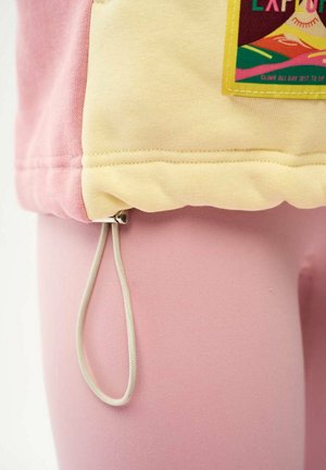 Pętla ze sznurka wisząca z dolnej krawędzi ubrania z różowym i żółtym materiałem oraz kolorową naszywką z tekstem.