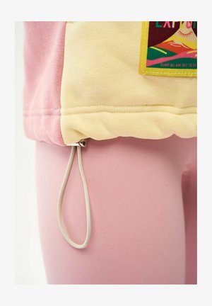 Pętla ze sznurka wisząca z dolnej krawędzi ubrania z różowym i żółtym materiałem oraz kolorową naszywką z tekstem.