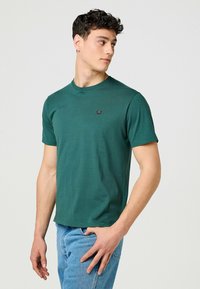 Grön t-shirt med kort ärm i bomull, med rund halsringning och en liten logotyp på bröstet. Buren med ljusblå jeans.