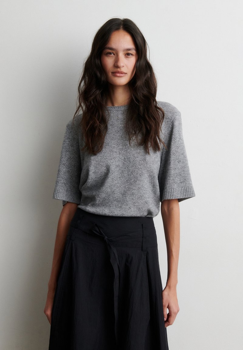Femme aux longs cheveux foncés portant un pull en maille gris à manches courtes et une jupe noire taille haute, debout devant un mur blanc uni.