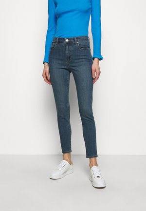 Persoon draagt slim-fit donkerblauwe jeans, felblauw lange mouwen shirt en witte sneakers, staand tegen een effen witte achtergrond.