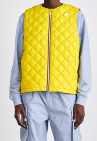 Persona che indossa un gilet trapuntato giallo con zip sopra una maglietta a maniche lunghe di colore azzurro chiaro e pantaloni cargo coordinati, in piedi contro uno sfondo semplice.