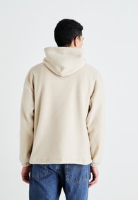 Beige hettegenser laget av mykt stoff, med en stor hette, ribbede mansjetter og en avslappet passform, kombinert med blå denimjeans.