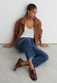 Kvinde med fletninger sidder på gulvet iført brun jakke, hvid tanktop, blå jeans og brune lædermokkasiner mod en hvid væg.
