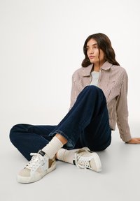 Randig avklippt jacka, grå tröja, mörkblå jeans, cremefärgade strumpor och vita sneakers med beige detaljer, sittande på en ljus bakgrund.