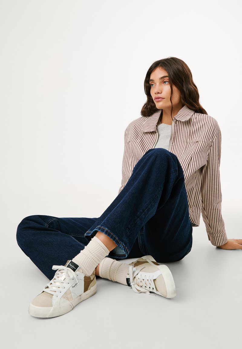 Randig avklippt jacka, grå tröja, mörkblå jeans, cremefärgade strumpor och vita sneakers med beige detaljer, sittande på en ljus bakgrund.