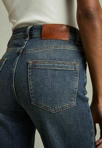 Jeans en denim bleu foncé à coupe droite, avec une patch en cuir marron portant l'inscription "REISS" et des poches arrière visibles avec des coutures contrastées.