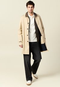 Beige regnrock med brun krage, knäppning framtill och ett enda innerfack. Bärs över vit polo och mörka jeans. Lätta skor och avslappnad passform.