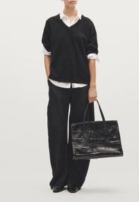 Maglione nero oversize con scollo a V, indossato sopra una camicia bianca. Pantaloni neri larghi e una borsa a tracolla in pelle nera testurizzata completano il look.