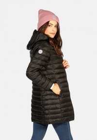 Doudoune noire avec un design matelassé, capuche et poches latérales. Portée avec un bonnet rose clair comportant un patch de logo rond.