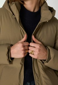 Lehká pufovaná bunda olivové barvy se zipovým zapínáním a kapucí. Má pružné manžety a pod ní je tmavý pletený svetr.