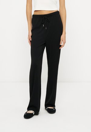 Femme portant un pantalon ample noir avec taille à cordon et des ballerines noires, debout devant un fond blanc.