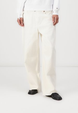 Personne portant un jean blanc ample avec des ourlets effilochés, associé à des chaussures noires à bout carré sur un fond blanc uni.