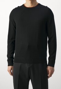 Homme portant un pull noir uni à manches longues et un pantalon noir, debout devant un fond blanc, les mains détendues le long du corps.