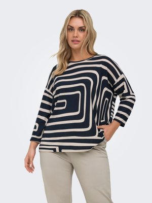 Femme aux longs cheveux blonds portant un haut à manches longues noir et beige à motif géométrique et un pantalon beige clair, debout avec une main dans la poche.