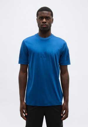 Man staat recht vooruit gericht, gekleed in een effen blauwe korte mouwen t-shirt en zwarte broek tegen een effen lichte achtergrond.