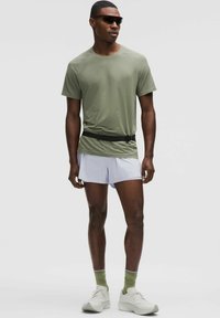 Short-sleeve olive green t-shirt with a rounded neckline, paired with white shorts, green-striped socks, and white sneakers. Black belt and sunglasses.