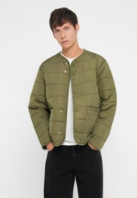 Quiltuota alyvuogių žalia striukė su apvaliu iškirptimu, spaudimo užtrauktukais ir dviem priekinėmis kišenėmis. Audinys lygus, su paminkštintu tekstūru.