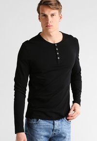 Camisa henley de manga larga negra, fabricada con un tejido suave, con un escote redondo y una tapeta de cinco botones en el pecho. Combinada con jeans azules.