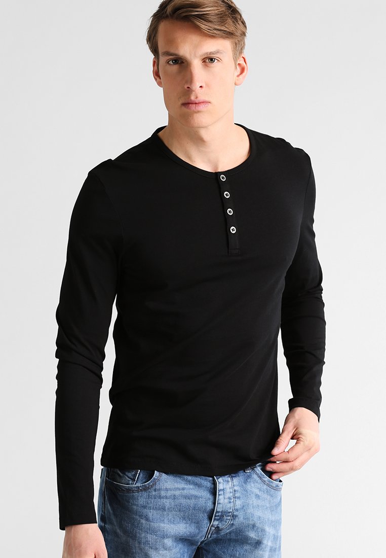Camisa henley de manga larga negra, fabricada con un tejido suave, con un escote redondo y una tapeta de cinco botones en el pecho. Combinada con jeans azules.