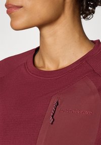 Haut de sport bourgogne en tissu texturé, col rond et manches raglan. Présente le logo Peak Performance près de l'épaule et un détail à cordon de serrage.