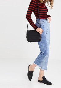 Borsa a tracolla nera con flap testurizzato, abbinata a una maglietta a righe rossa e nera a maniche lunghe, jeans chiari sfilacciati e scarpe slip-on nere.