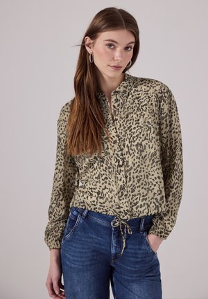 Chemise boutonnée en imprimé léopard beige et noir, en tissu léger, manches longues avec poignets élastiques, taille nouée, associée à un jean bleu.