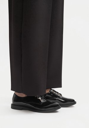 Czarne buty ze skóry lakierowanej o zaokrąglonym nosku, wiązane na sznurówki z złotymi przelotkami, w połączeniu z luźnymi czarnymi spodniami.