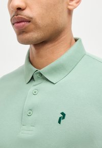 Gros plan d'une personne portant un polo vert clair avec un col, trois boutons et un petit logo brodé vert foncé sur la poitrine.